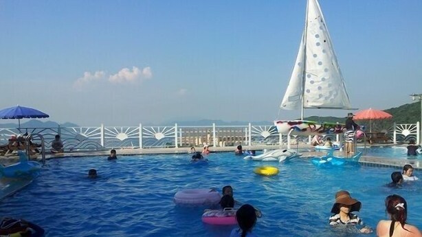 A swimming pool with multiple people, some on inflatable rings, and a sailboat in the background.