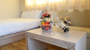 A hotel room with a bed, a small table with a fruit basket, two wine glasses, and a teddy bear.