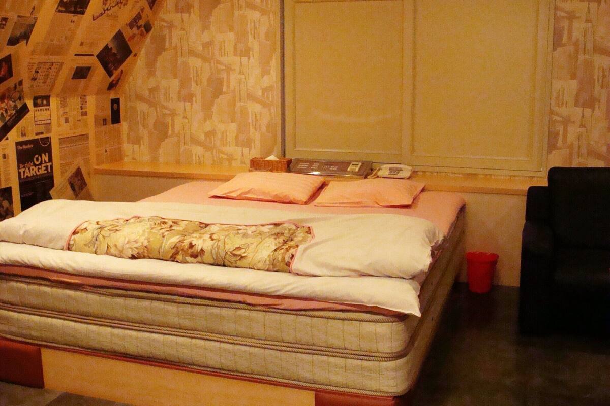 A single bed with a floral-patterned blanket and pillows, a bedside table with a lamp, and a red trash bin.