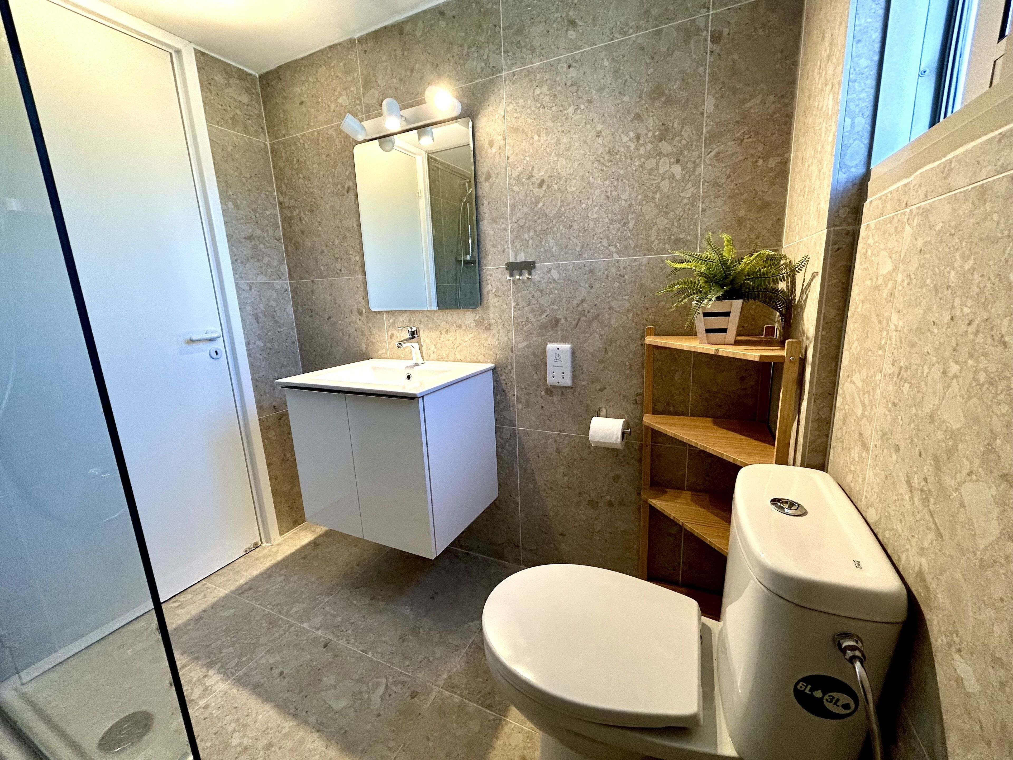 Un baño moderno con lavamanos, espejo y retrete blancos. Hay una planta sobre una repisa y una ducha visible al fondo.