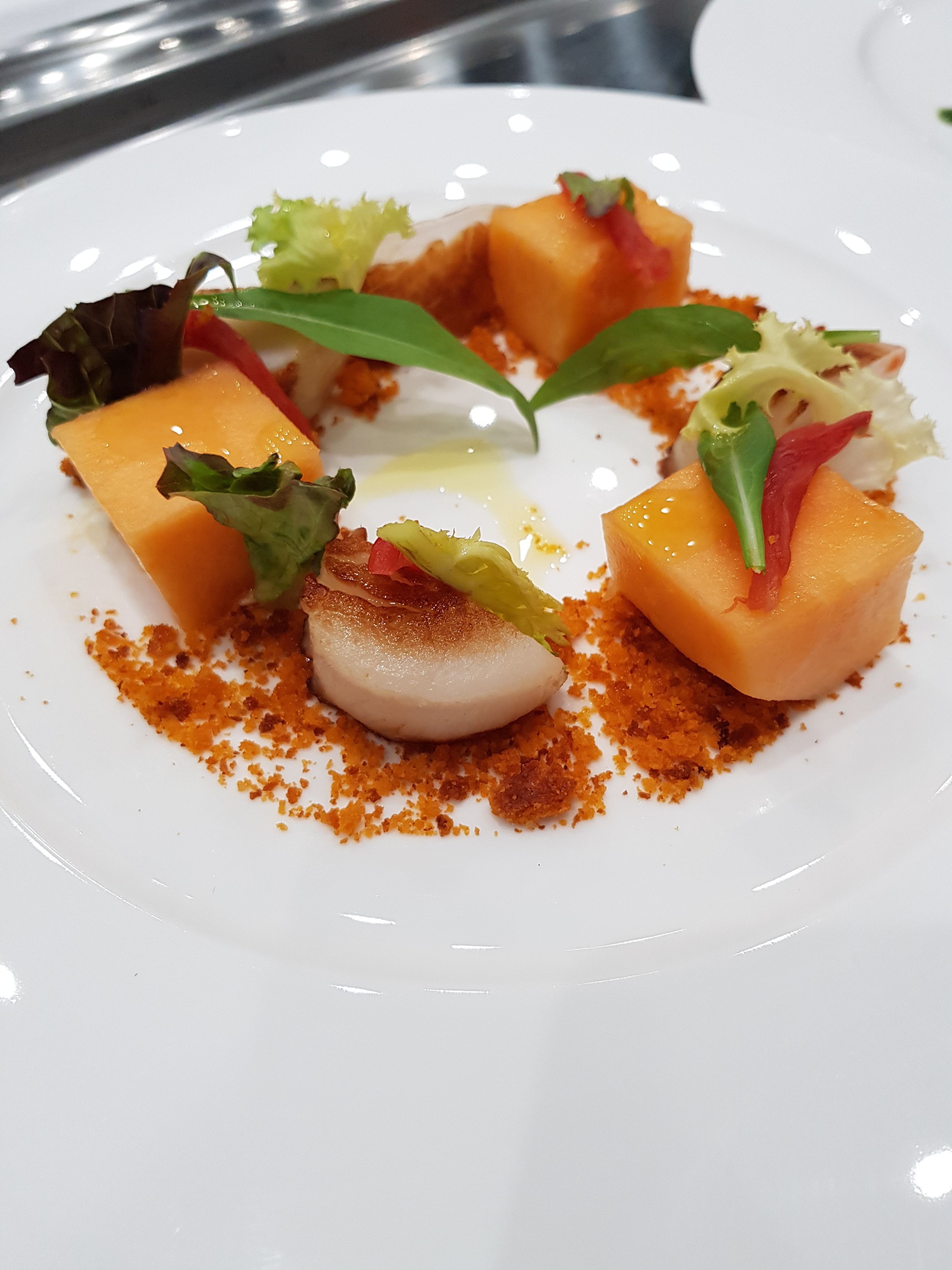A white plate with a dessert consisting of a custard-like sphere, cubes of orange fruit, and green leaves.