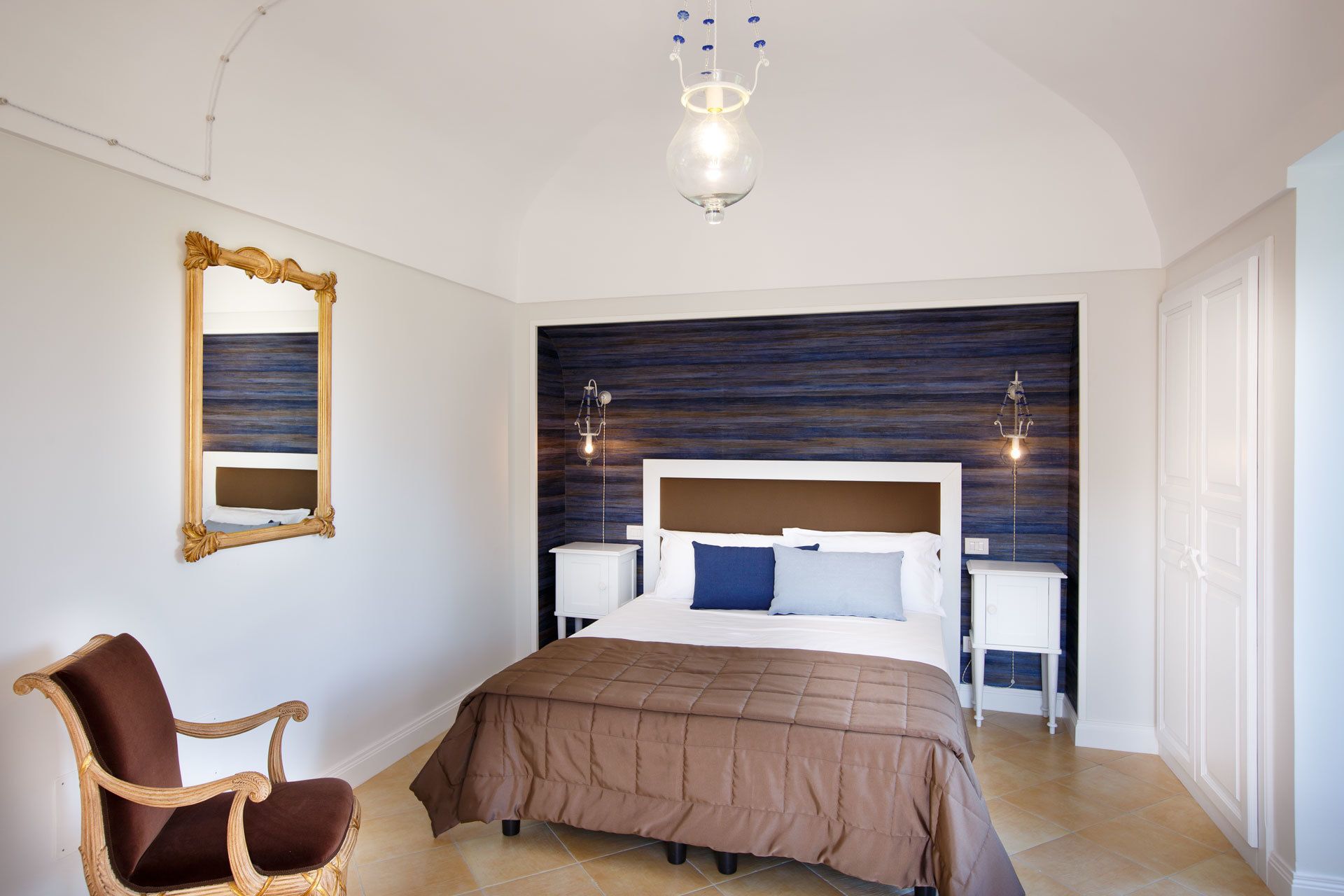 A bedroom with a large bed, a nightstand, a chair, a mirror, and a chandelier.
