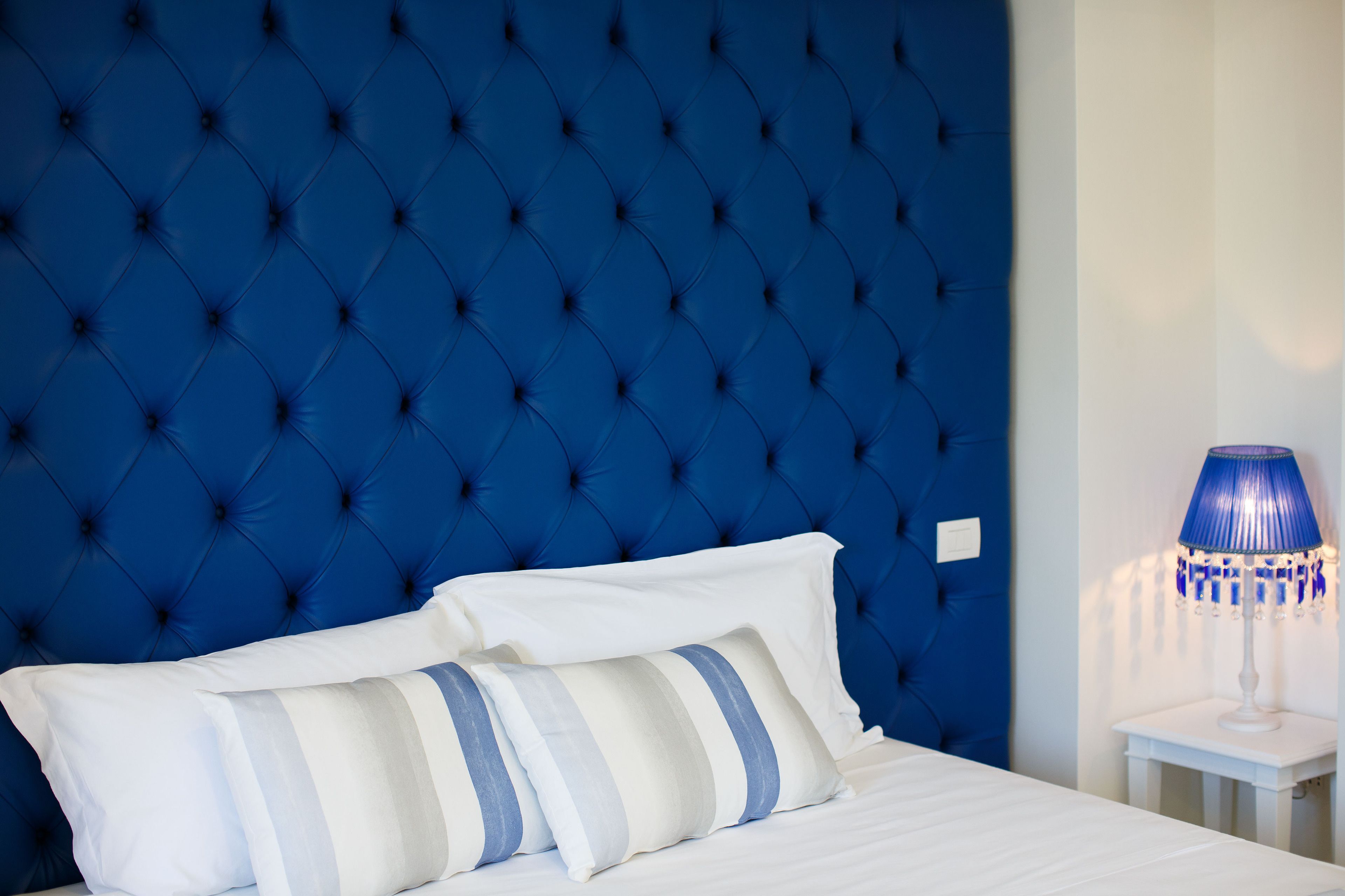 Un letto con cuscini bianchi e a righe, una testata imbottita blu e un comodino bianco con una lampada blu.