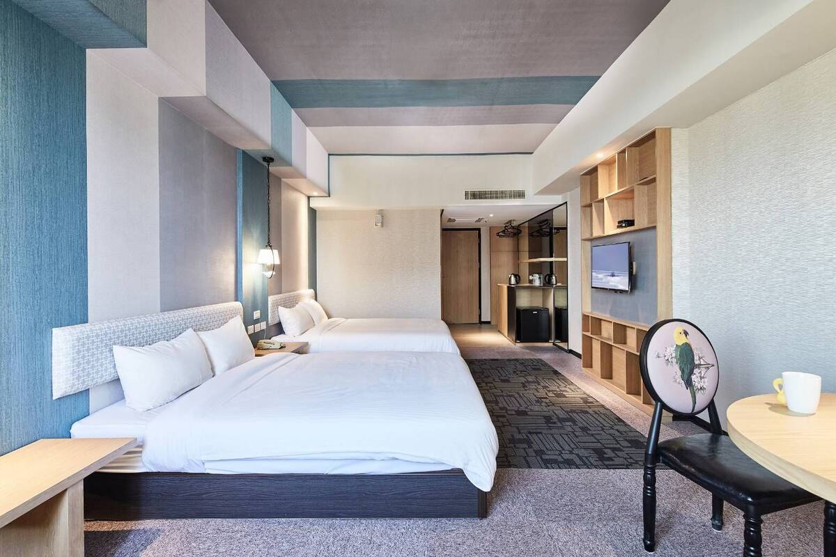 A modern hotel room with two beds, a desk, a chair, a TV, and a small table.