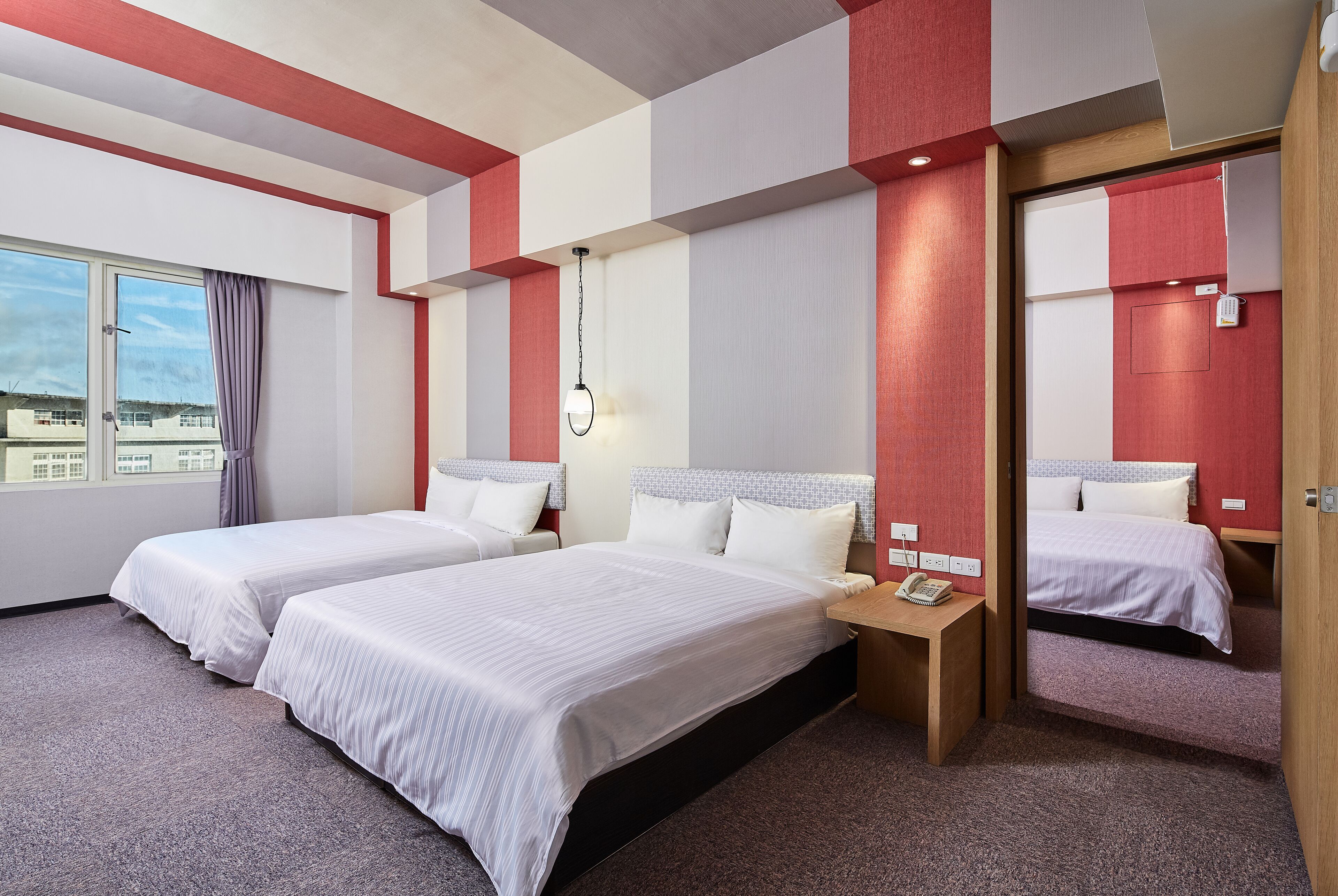 A hotel room with two beds, a window with curtains, a bedside table with a phone, and a wall-mounted light fixture.