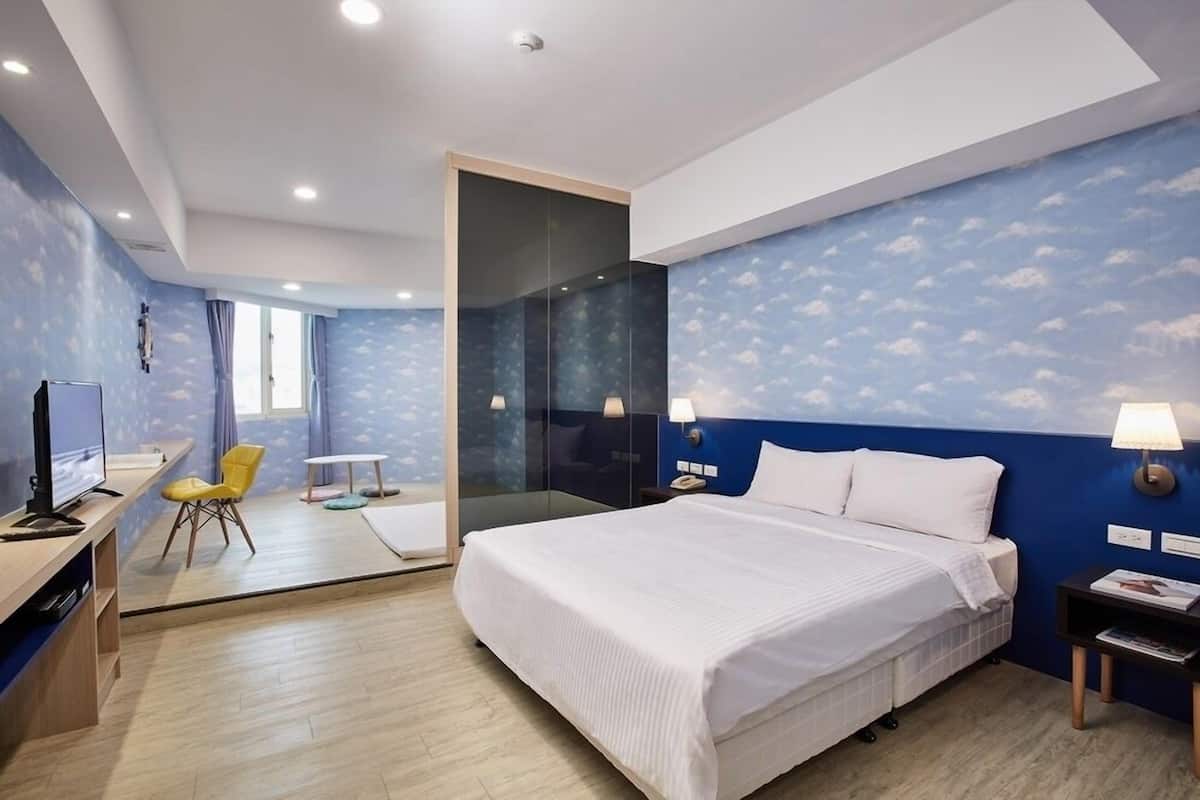 A modern hotel room with a large bed, a desk with a computer, a chair, and a glass partition separating the room from another space.