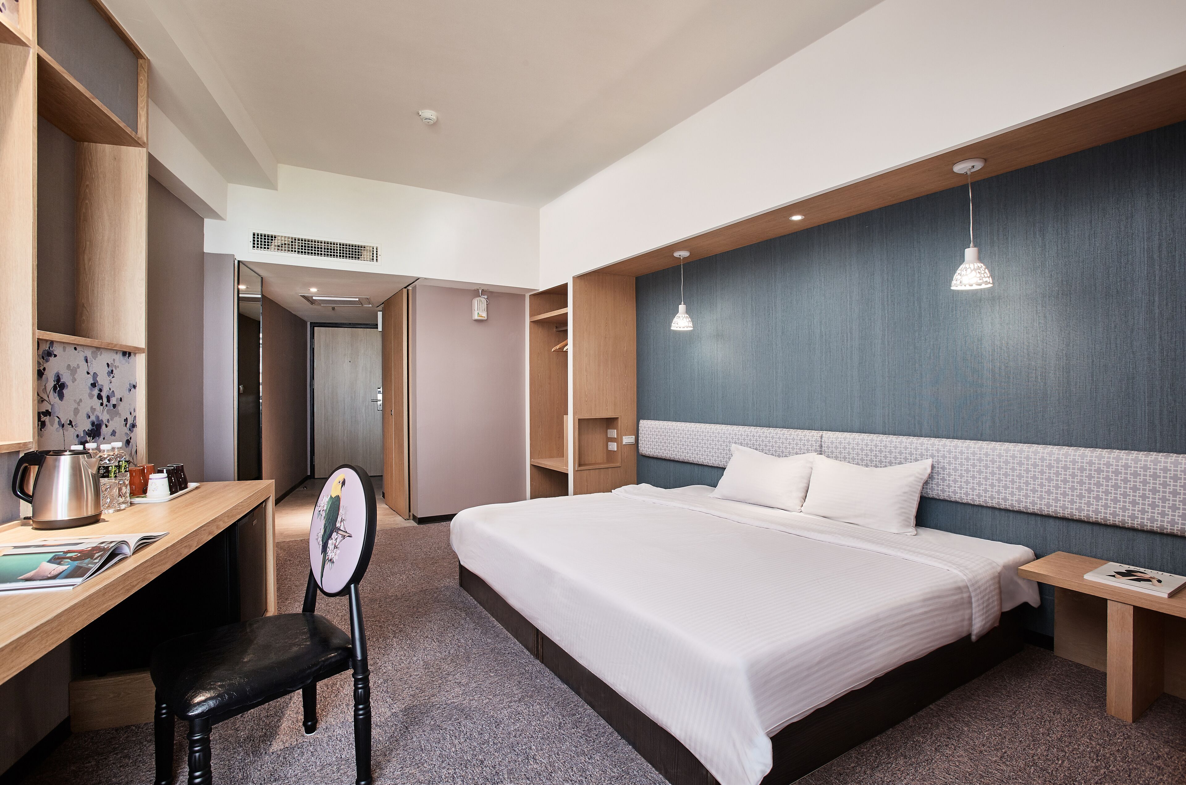 A modern hotel room with a large bed, a desk with a chair, and a small table with a lamp.