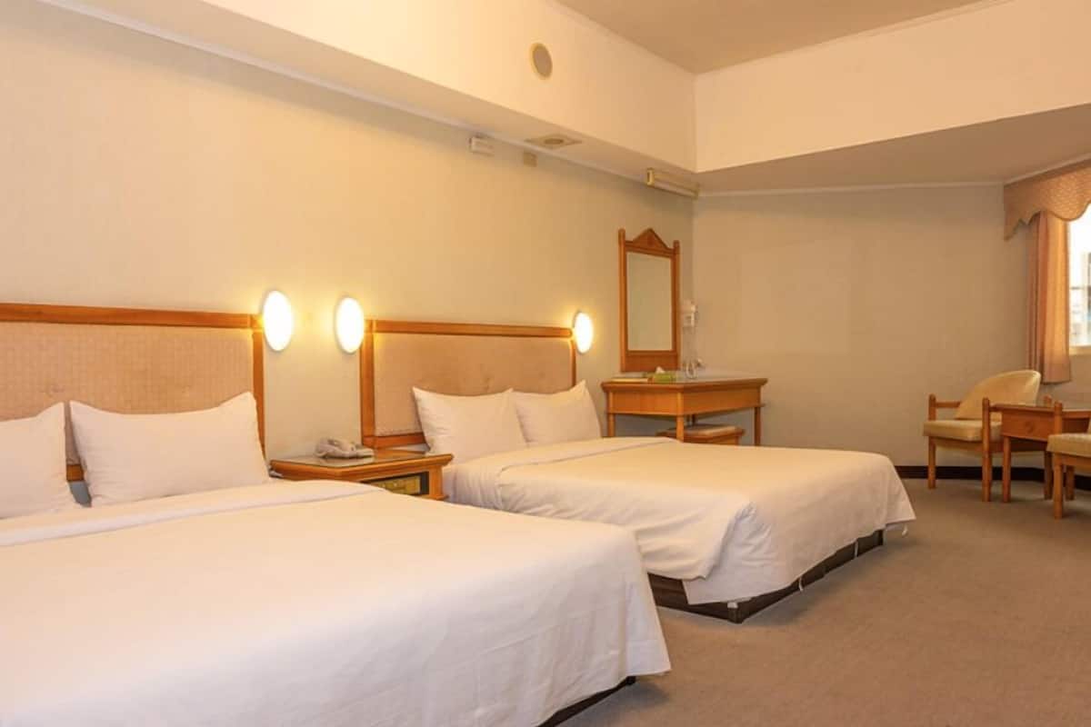 A hotel room with two single beds, a wooden headboard, bedside tables with lamps, a mirror, a chair, and a desk.