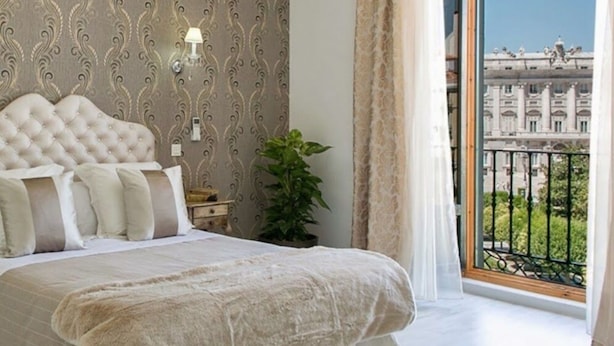A bedroom with a large bed, a wall clock, a bedside table, a potted plant, and a view of a building through a balcony. - Hostal Central Palace Madrid (Madrid)