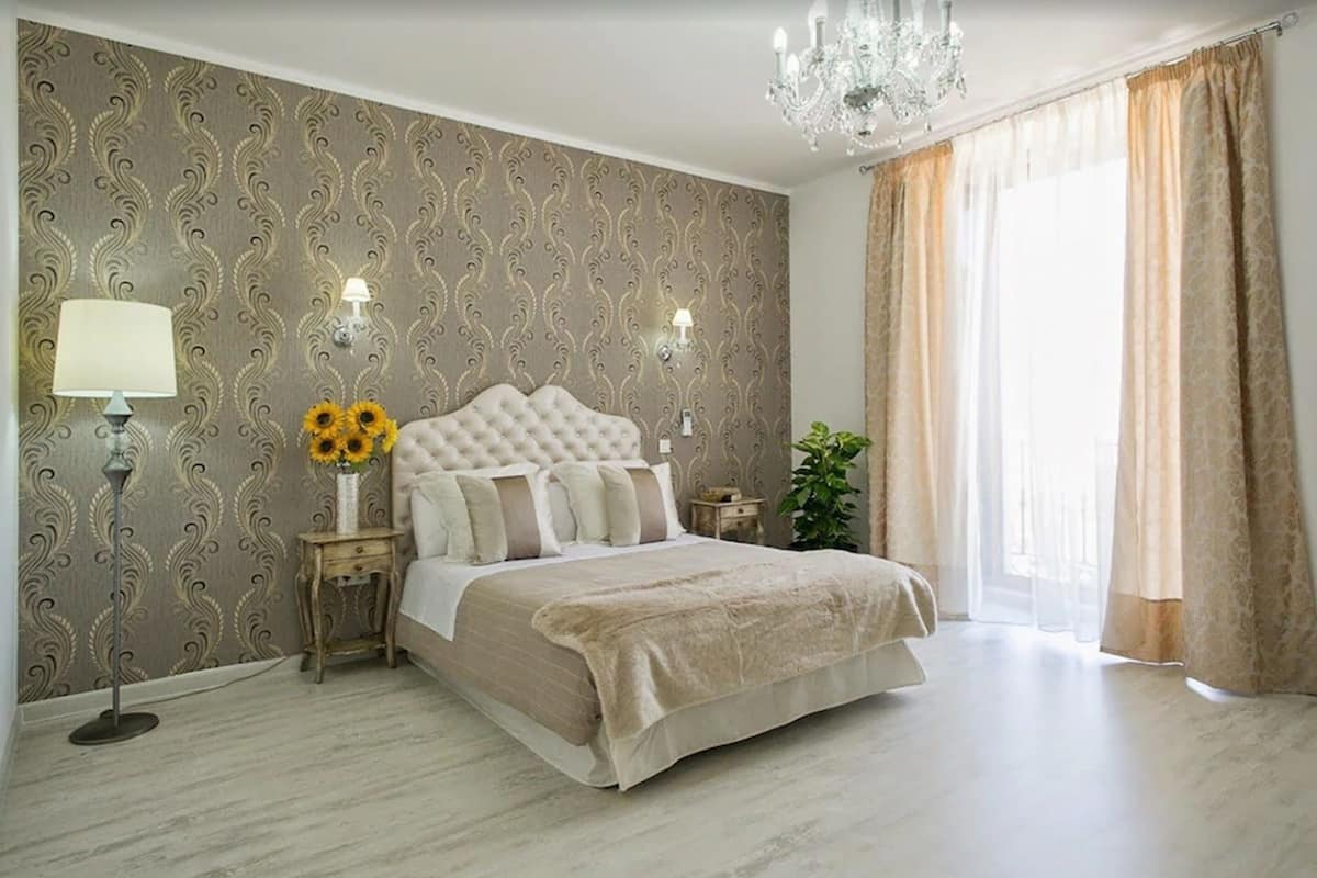 Un dormitorio con cabecera capitonada, dos lámparas de noche, una mesita de noche con un jarrón de girasoles y una ventana grande con cortinas.