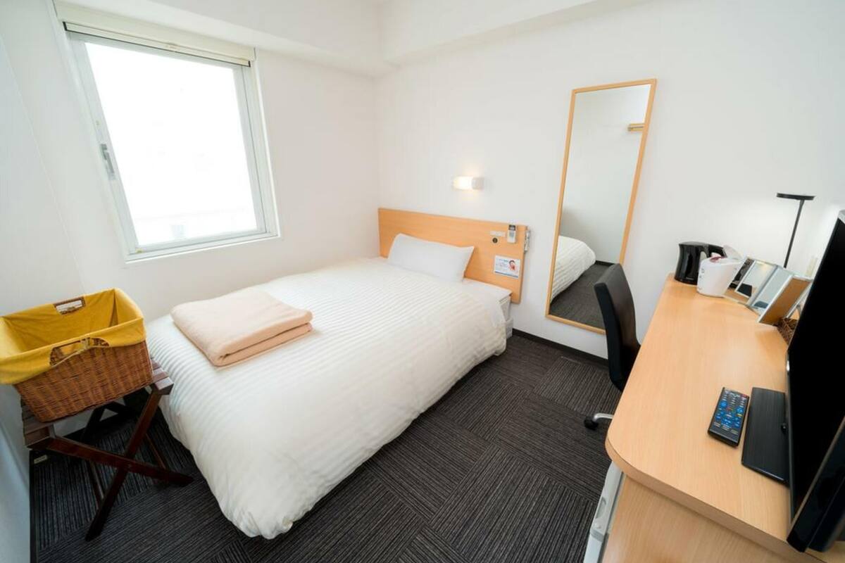 Ein Hotelzimmer mit Bett, Schreibtisch, Stuhl, Spiegel, Fenster und Mülleimer.