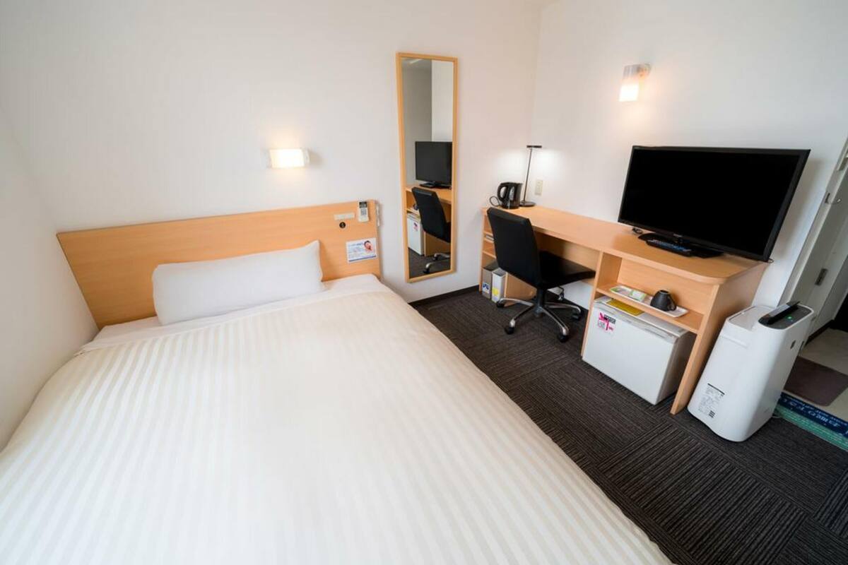 Ein Hotelzimmer mit Bett, Schreibtisch mit Computer, Fernseher, Spiegel und einer kleinen Küchenzeile.