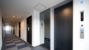 Ein Hotelgang mit Schachbrettmuster-Teppich, Türen und einem Aufzug mit digitaler Anzeige.