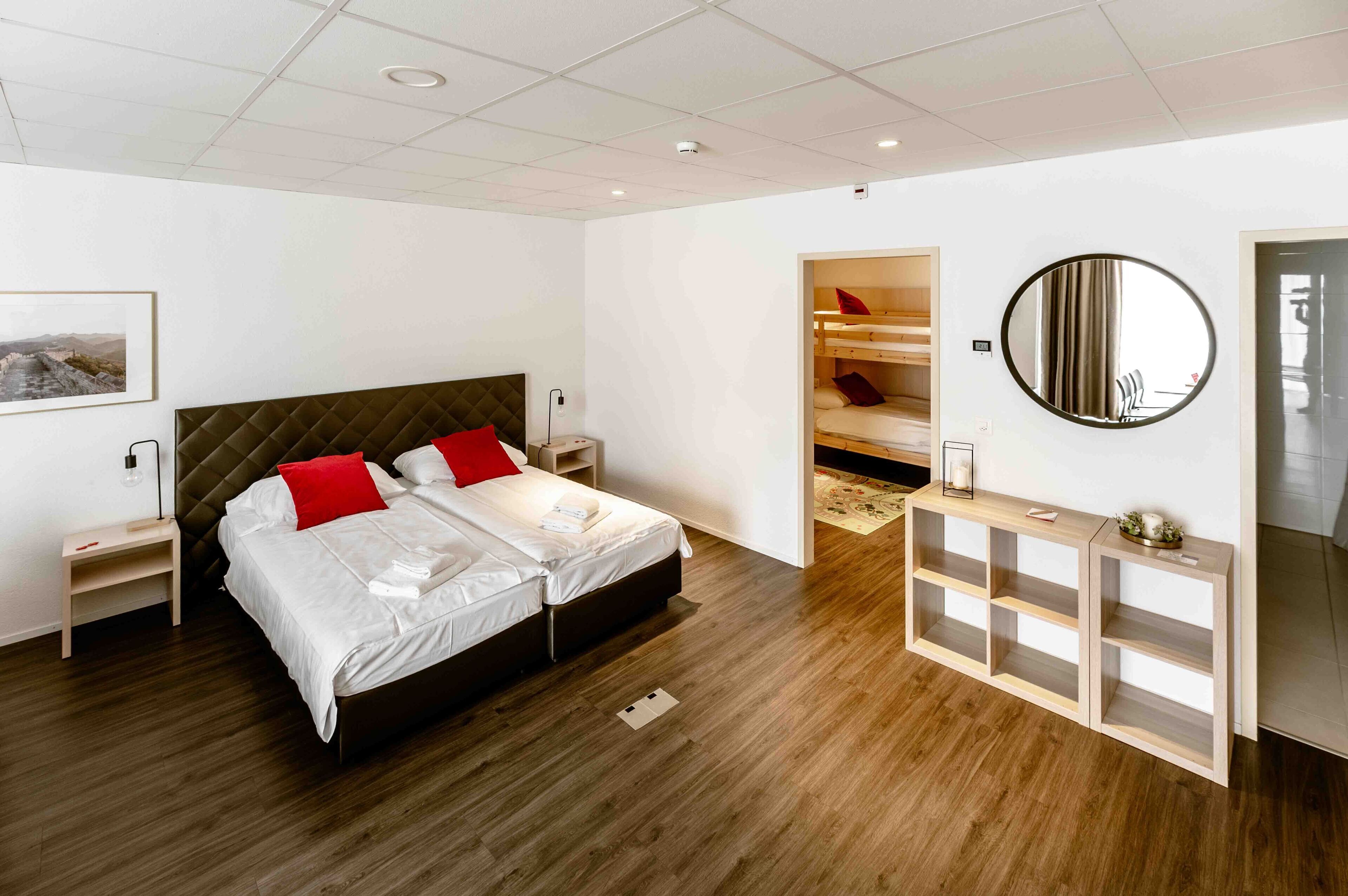 A modern hotel room with a large bed, bedside tables, a wooden console, a round mirror, and a built-in wardrobe.