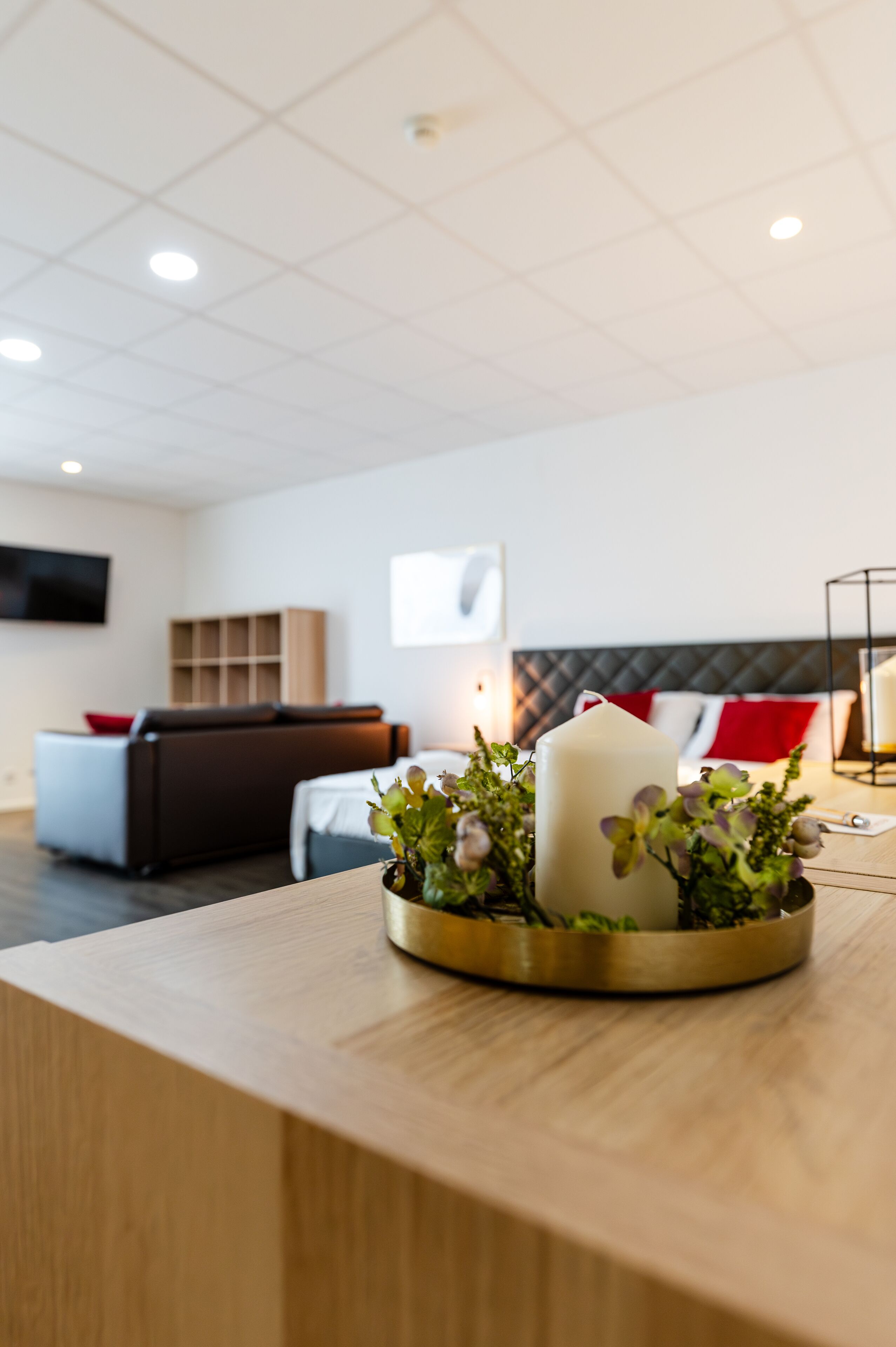 A modern hotel room with a wooden reception desk, a large white candle centerpiece, a sofa with red cushions, and a flat-screen TV mounted on the wall.