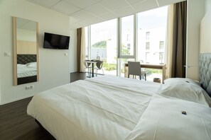 Une chambre d’hôtel dotée d’un grand lit, d’un bureau avec une chaise, d’une télévision et d’un balcon offrant une vue sur les bâtiments et la verdure.