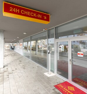 L’entrée d’un hôtel, avec une pancarte indiquant un service d’enregistrement ouvert 24 heures sur 24, des portes en verre et un tapis rouge orné d’un texte.