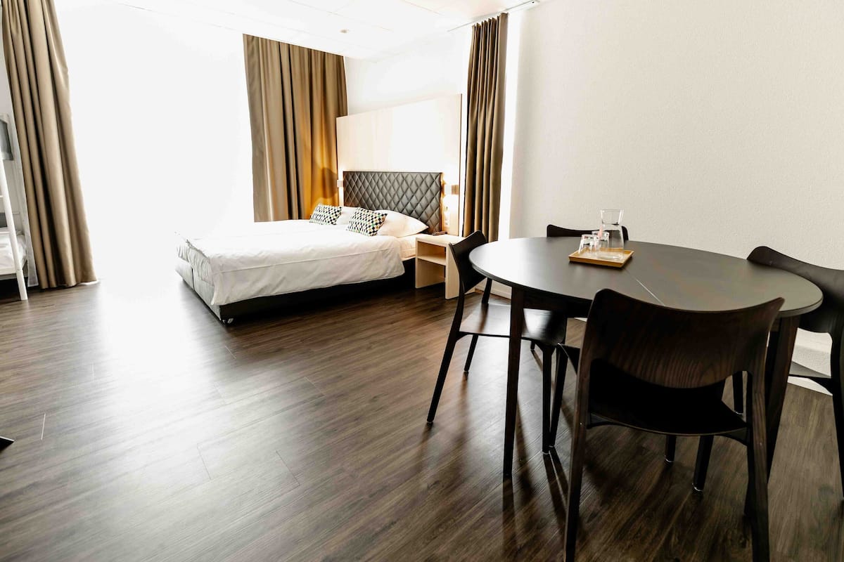Hotellihuone, jossa on sänky, yöpöytä, lamppu ja pyöreä ruokapöytä tuoleineen.