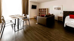 A modern hotel room with a dining area, a sofa, a bed, a TV, and a bookshelf.