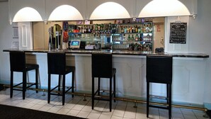 Una zona de bar con tres taburetes negros, un mostrador con una caja registradora y estanterías llenas de botellas y vasos.