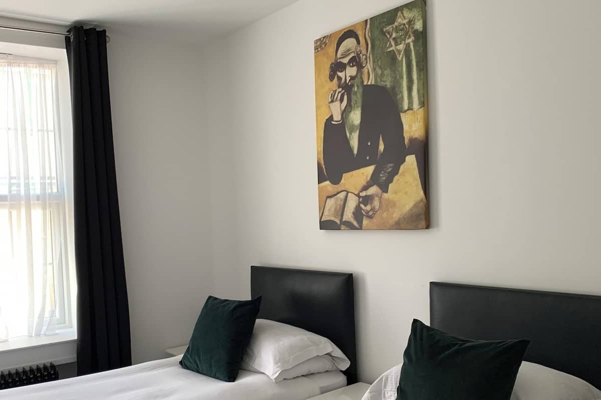 Habitación de hotel con dos camas, cada una con cabecero oscuro y ropa de cama blanca. Hay un cuadro en la pared sobre las camas y una ventana con cortinas al lado izquierdo.