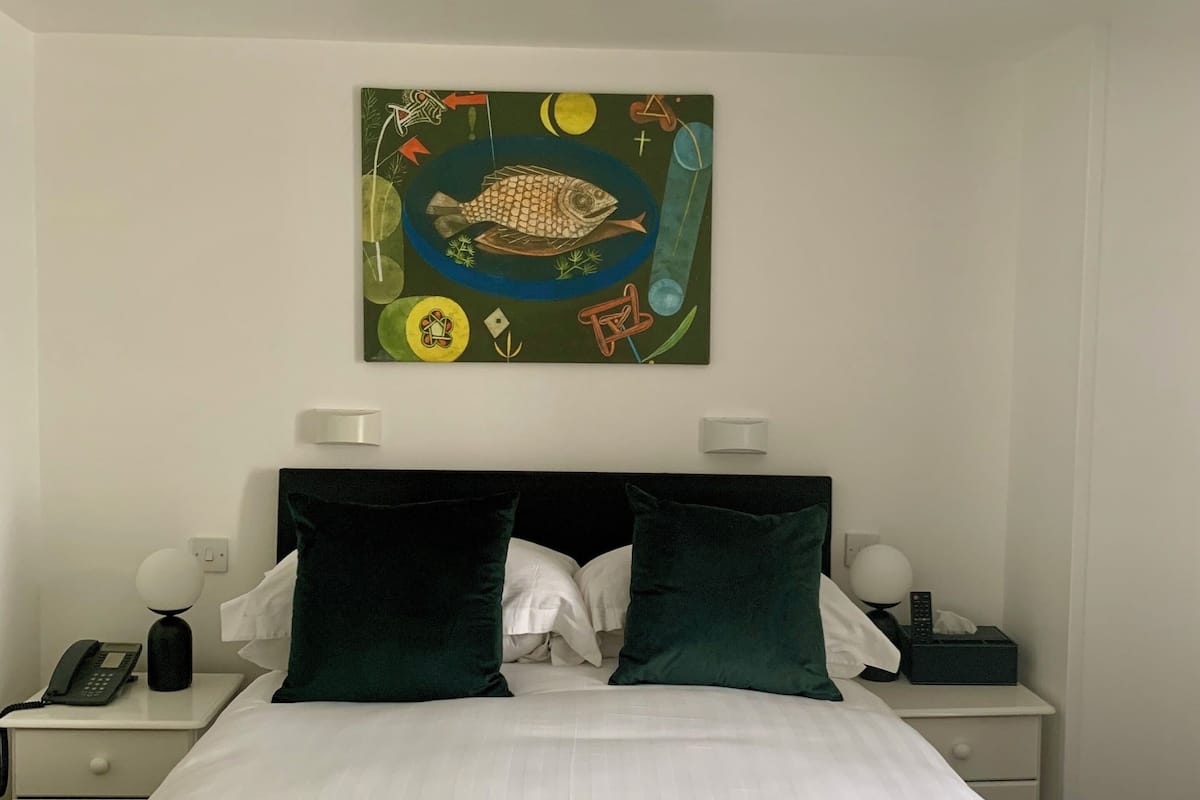 Una cama bien tendida con sábanas blancas, dos almohadas verde oscuro y una almohada blanca. Encima de la cama hay un cuadro abstracto y colorido.