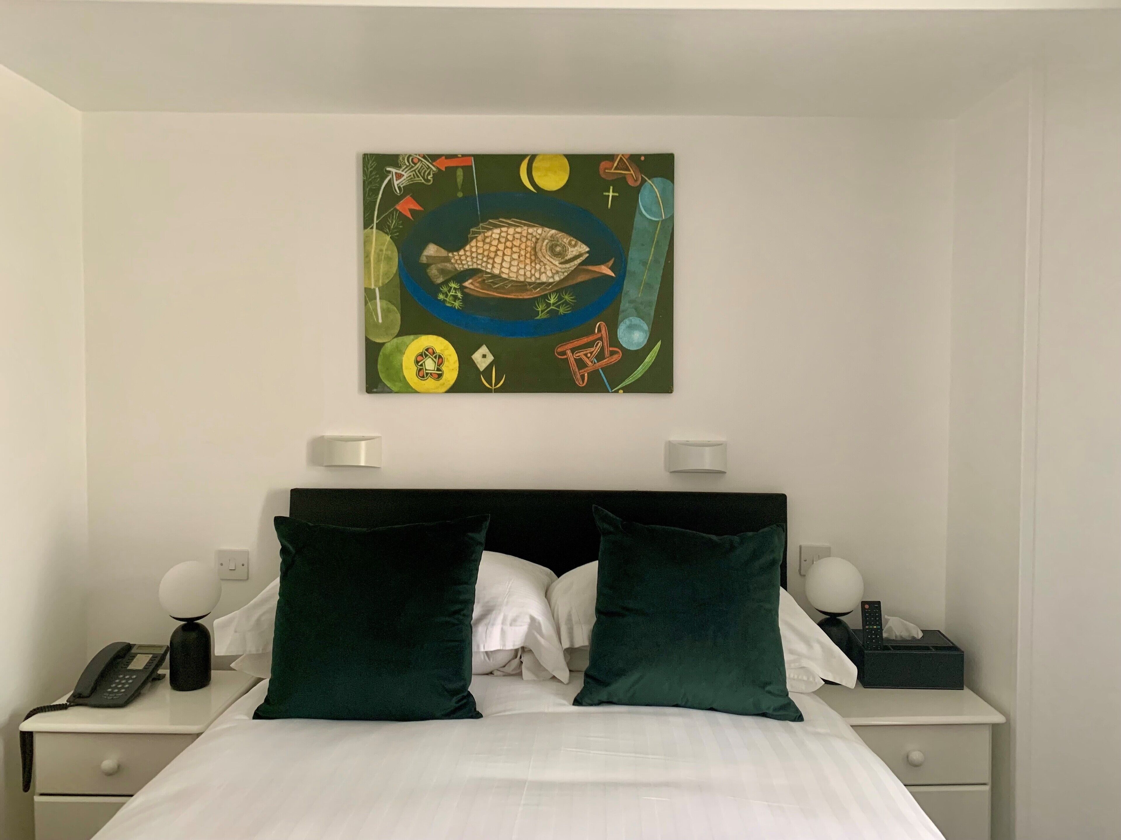 A hotel room with a bed, two bedside tables, a nightstand, a wall-mounted artwork, and a telephone.