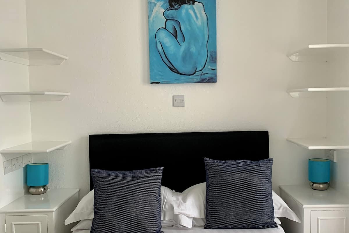 Un dormitorio con cama, dos almohadas, mesita de noche, lámpara y un cuadro en la pared.