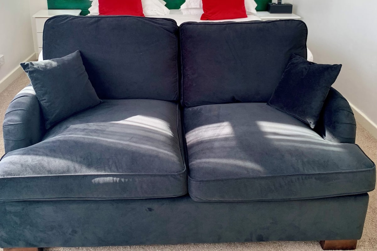 Una sala de estar moderna con un sofá gris oscuro, almohadas rojas y blancas, y un cabecero verde. Un cuadro abstracto y colorido adorna la pared sobre el sofá.