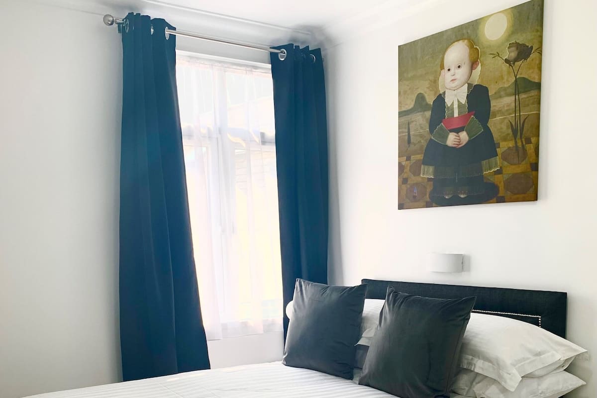 Una cama bien tendida con ropa de cama blanca y almohadas gris oscuro, una mesita de noche con una lámpara, y un cuadro de una niña en la pared.