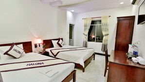 Quarto de hotel com três camas identificadas como 'HALONA', uma escrivaninha de madeira, uma cadeira, televisão fixada na parede e janela com cortinas.