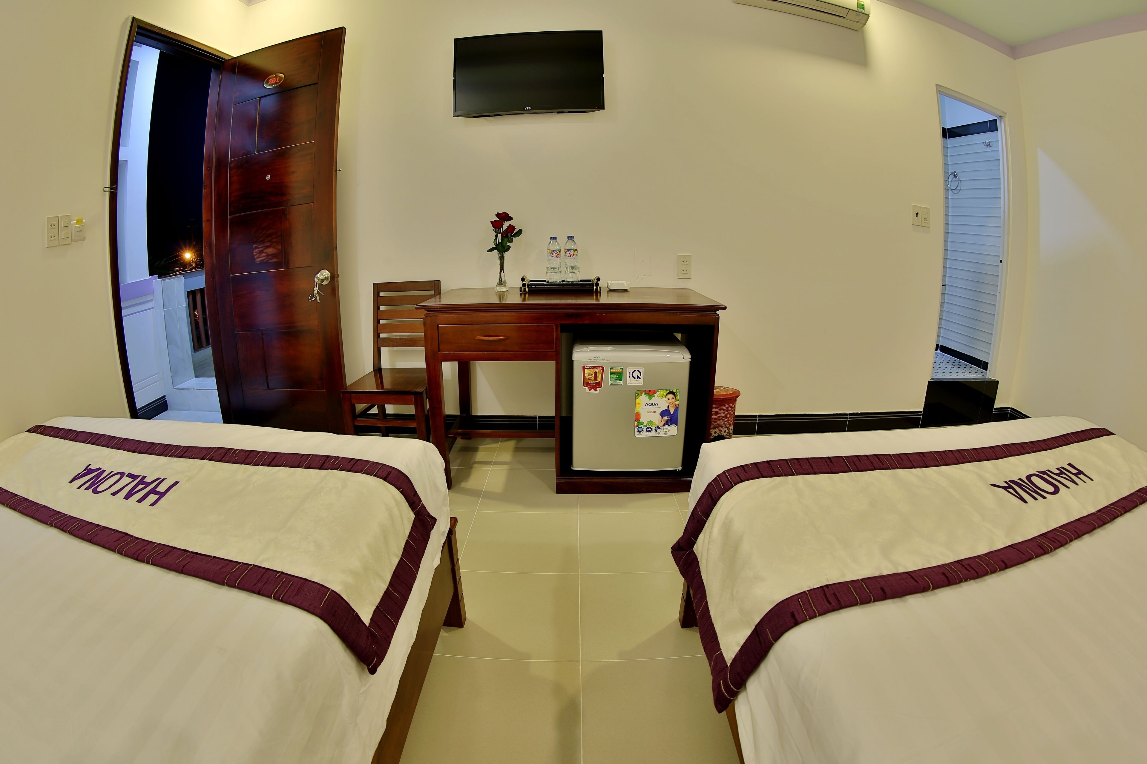 Habitación de hotel con dos camas identificadas con la etiqueta "HALONA", un escritorio con refrigerador, un televisor en la pared y una puerta que da a otra habitación.