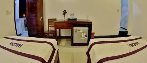 Habitación de hotel con dos camas identificadas con la etiqueta "HALONA", un escritorio con refrigerador, un televisor en la pared y una puerta que da a otra habitación.