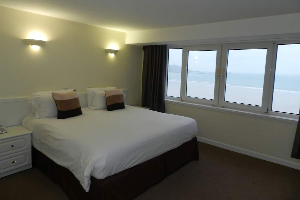 A hotel room with a large bed, two bedside lamps, a nightstand, and a window with a view of the beach.