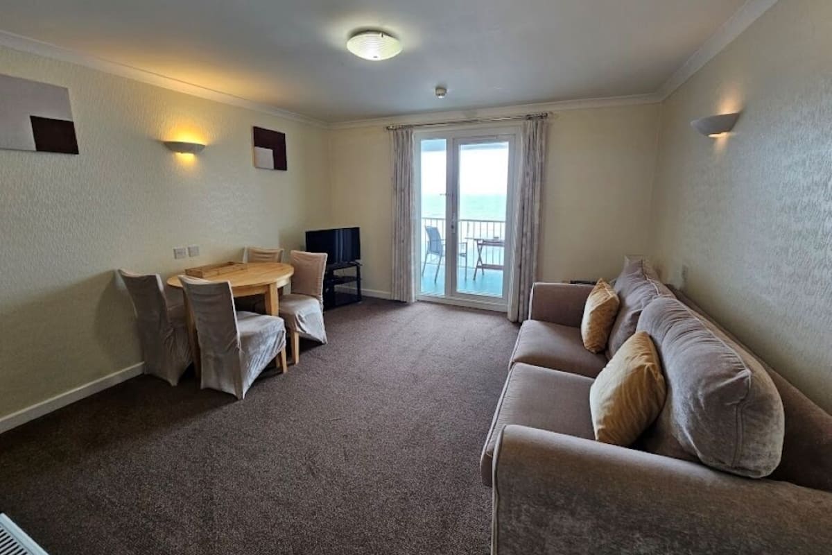 A hotel room with a sofa, a dining table with chairs, a TV, and a view of the sea through the windows.
