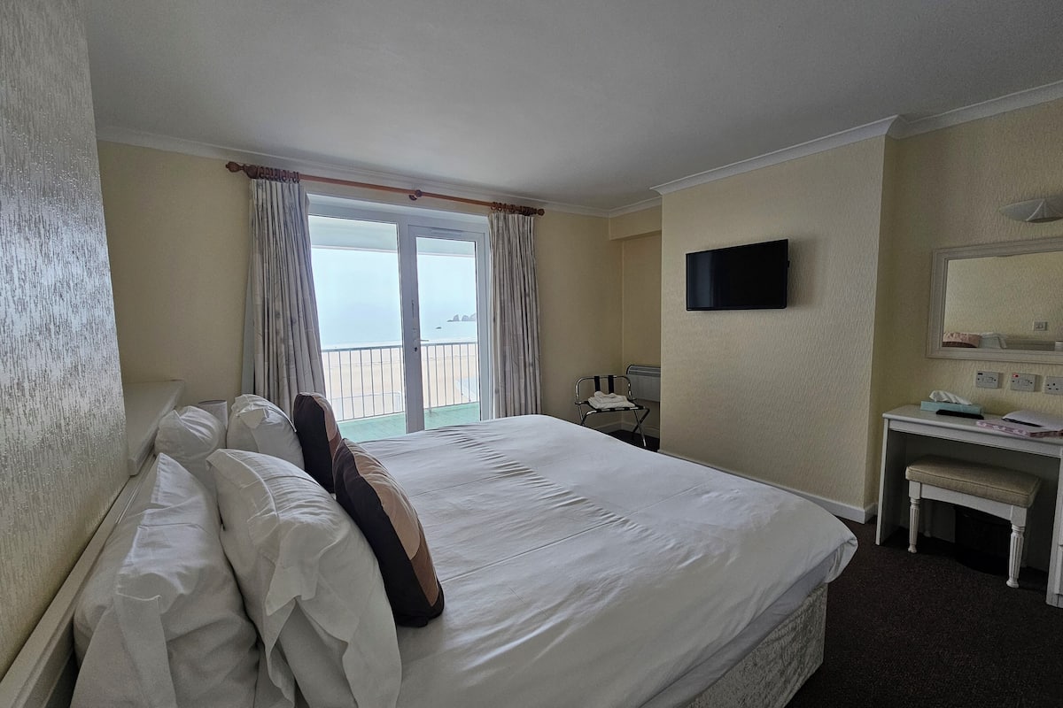A hotel room with a large bed, a desk, a chair, a TV, and a balcony with a view of the sea.