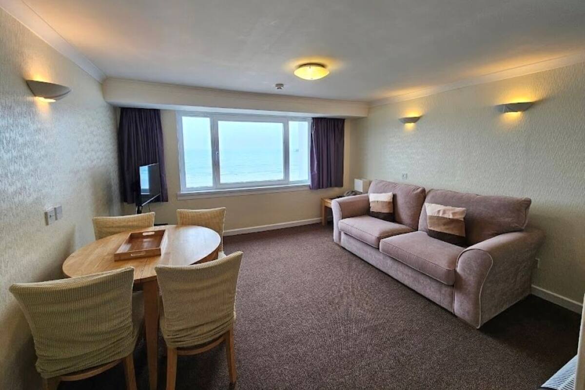 A compact living room with a round wooden table, four wicker chairs, a beige sofa, and a large window with curtains.