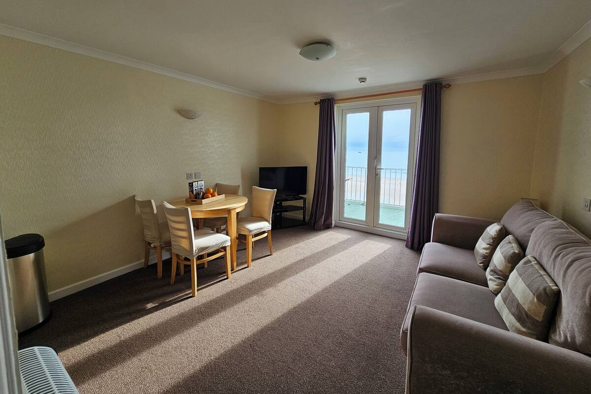 A hotel room with a dining area, a sofa, a TV, and a balcony with a view of the beach.