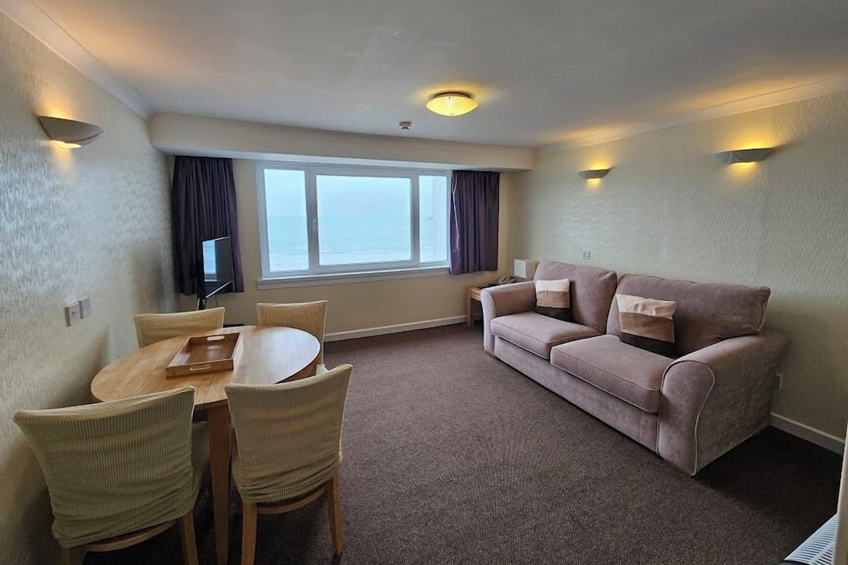 A room with a dining table, chairs, a sofa, a TV, and a window with curtains.