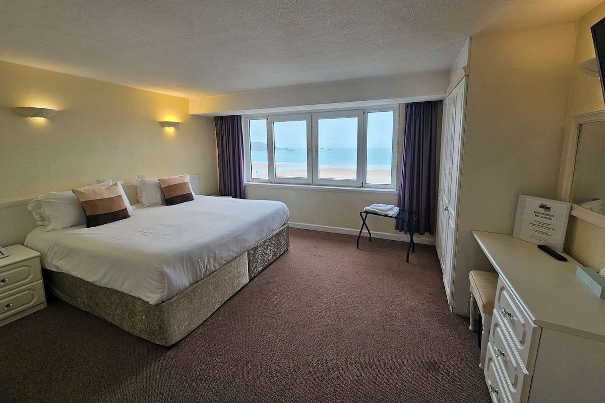 A hotel room with a large bed, a bedside table, a desk with drawers, a chair, a window with a view of the beach, and a wall-mounted light fixture.