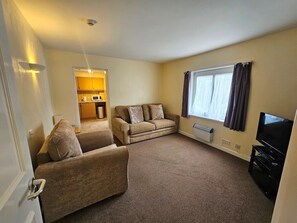 A living room with a sofa, a television, and a window with curtains. - The Beau Rivage (St. Brelade)