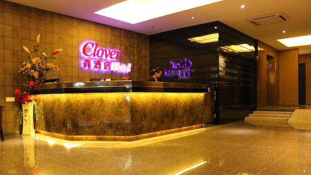A hotel reception area with a marble counter, a neon sign, and a staircase leading to the upper floor. - Clover Hotel (Masai)