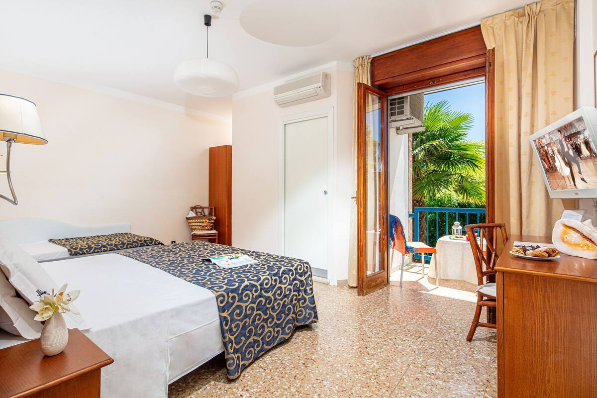 A hotel room with a bed, bedside table, lamp, and a view of the outside balcony.