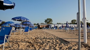 Una playa con hileras de sillas y sombrillas azules, un paseo marítimo arenoso y un edificio al fondo.