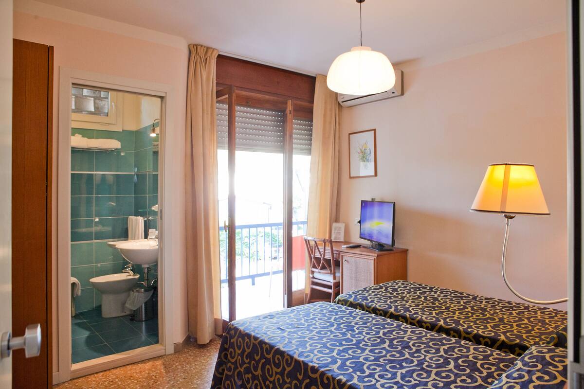 A hotel room with a bathroom, two beds with patterned bedspreads, a desk with a computer, a lamp, and a window with curtains.