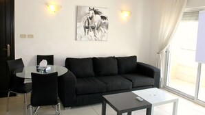 Ein modernes Wohnzimmer mit einem schwarzen Sofa, einem Glastisch und einem runden Esstisch mit Stühlen. An der Wand hängt ein Bild von Pferden und zwei Wandleuchten.