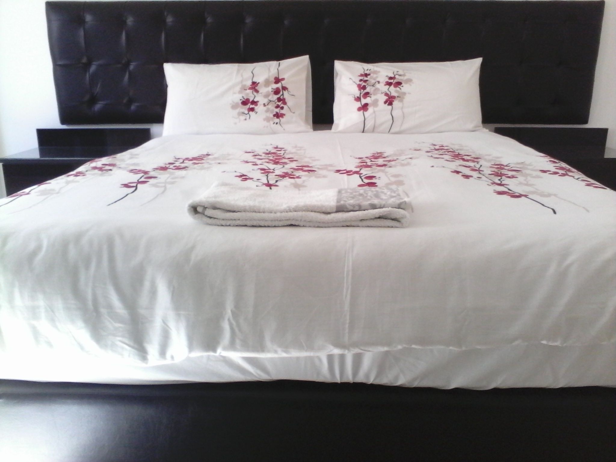 A bed with a white bedspread and two pillows with floral designs, a folded white towel, and a dark headboard.