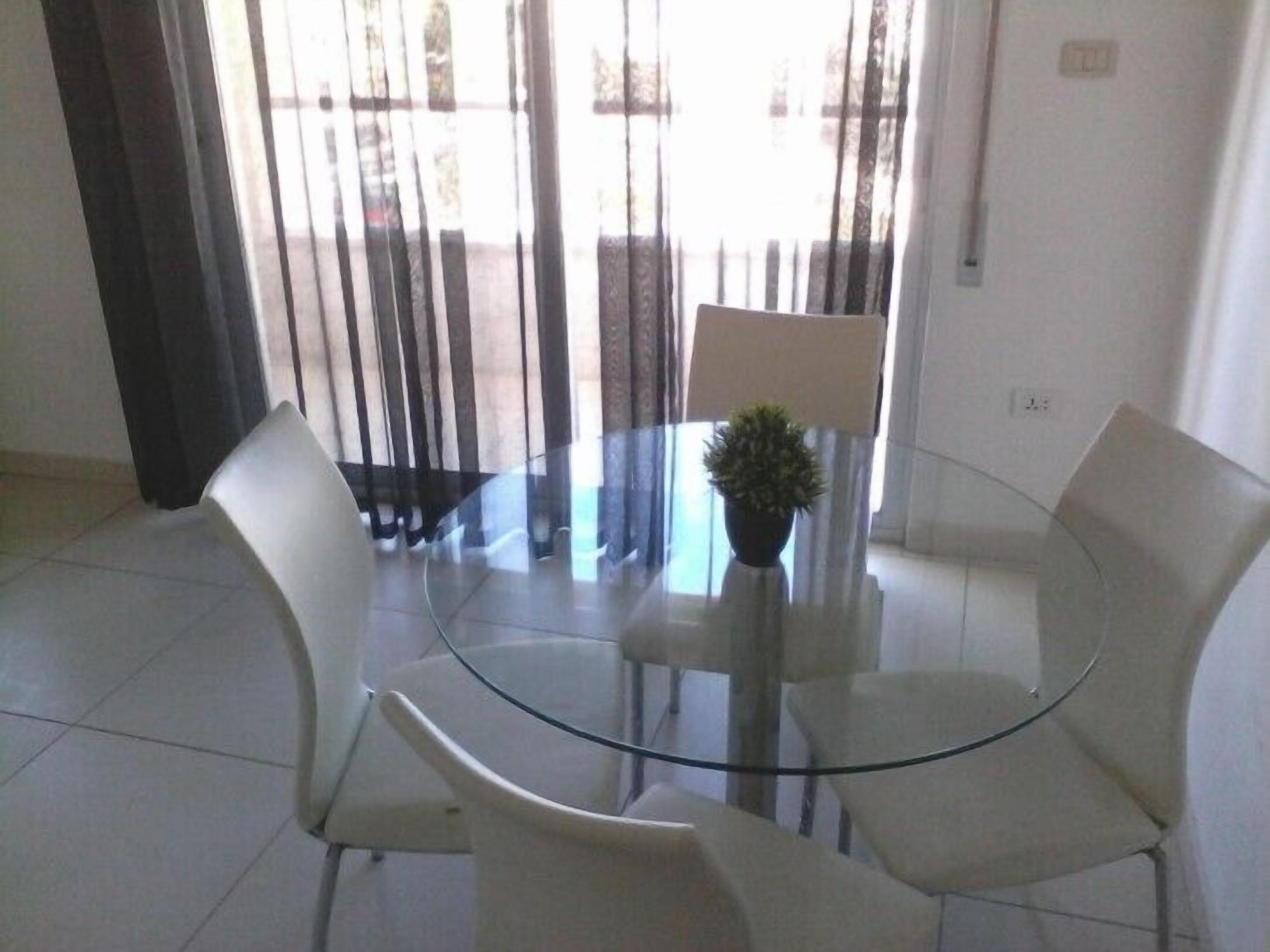 Un espace repas moderne avec une table en verre et des chaises blanches, une plante en pot et une vue à travers une fenêtre équipée de stores.