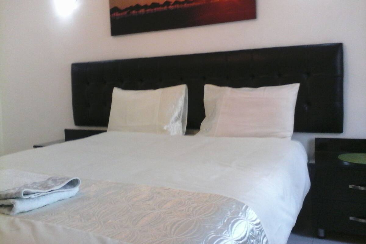 A neatly made bed with white linens and pillows, a black headboard, and a painting of a sunset on the wall.