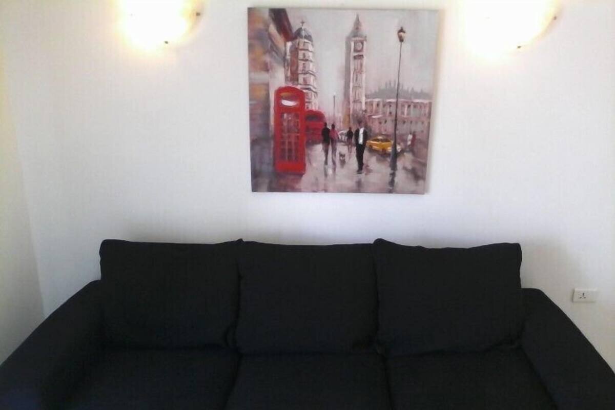 A living room with a black sofa, a painting of a cityscape with a red telephone booth, and a white wall with a light switch.